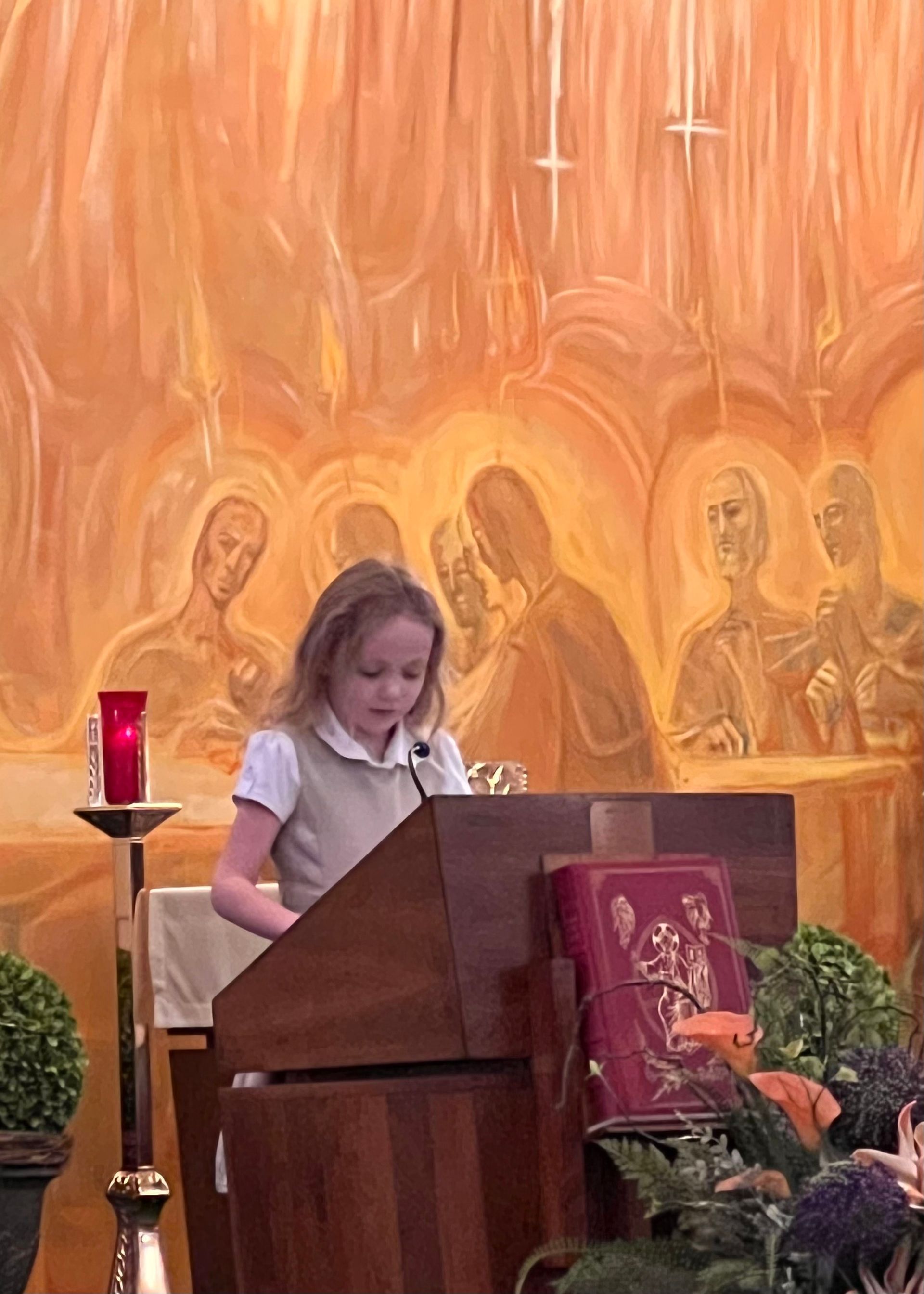 Young girl in school uniform reads at a podium in front of a religious painting.
