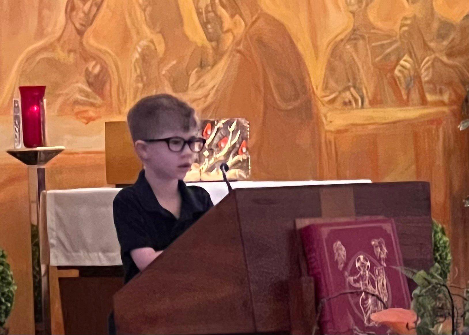 Young boy with glasses speaking at a church lectern; an ornate mural is in the background.