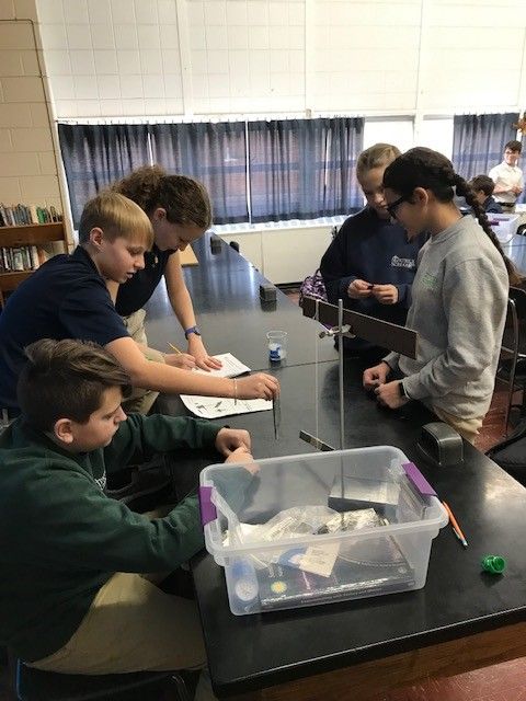 Students in a classroom conduct a science experiment. They lean over a table with materials, focused.