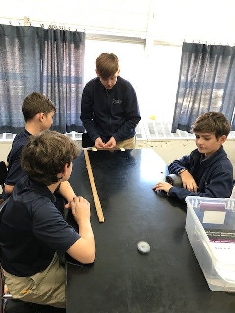 Four boys in a classroom at a table experimenting with objects.