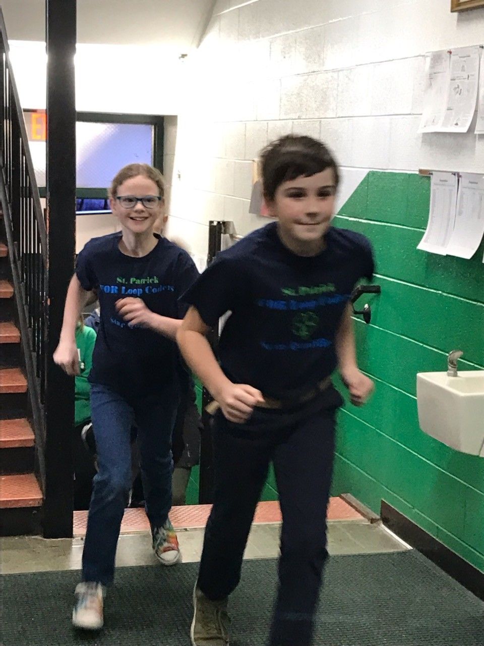 Two children in matching blue shirts run down a stairwell, smiling.