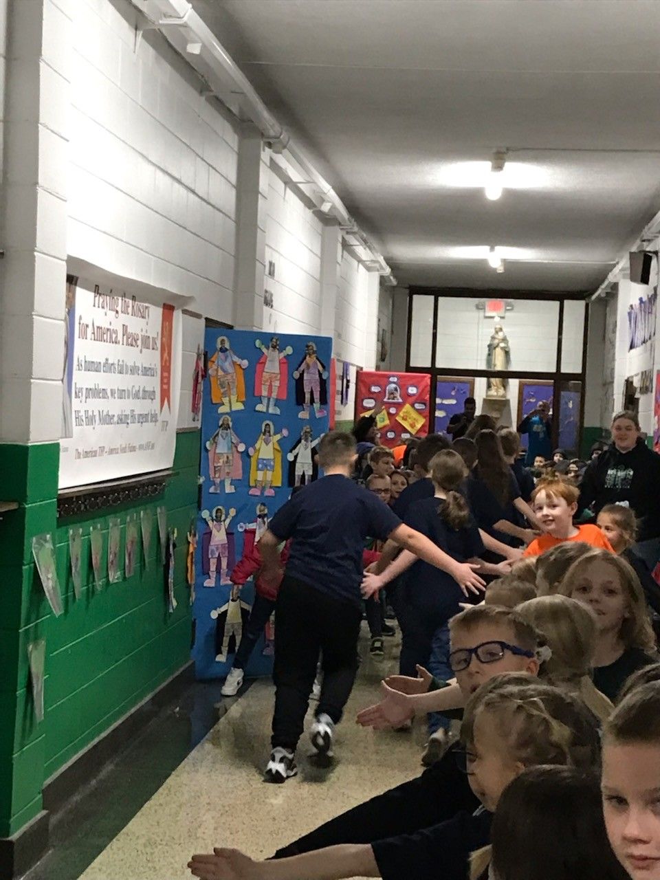 Children walking in a school hallway, some reaching out, with decorations on the wall.