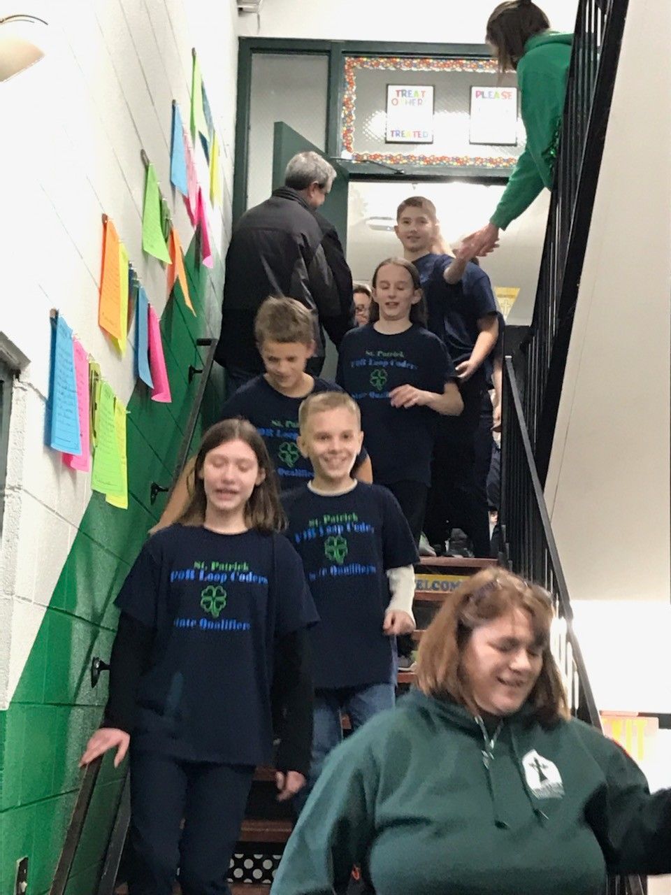 Children in matching shirts walk down school stairs, a teacher in green assists.