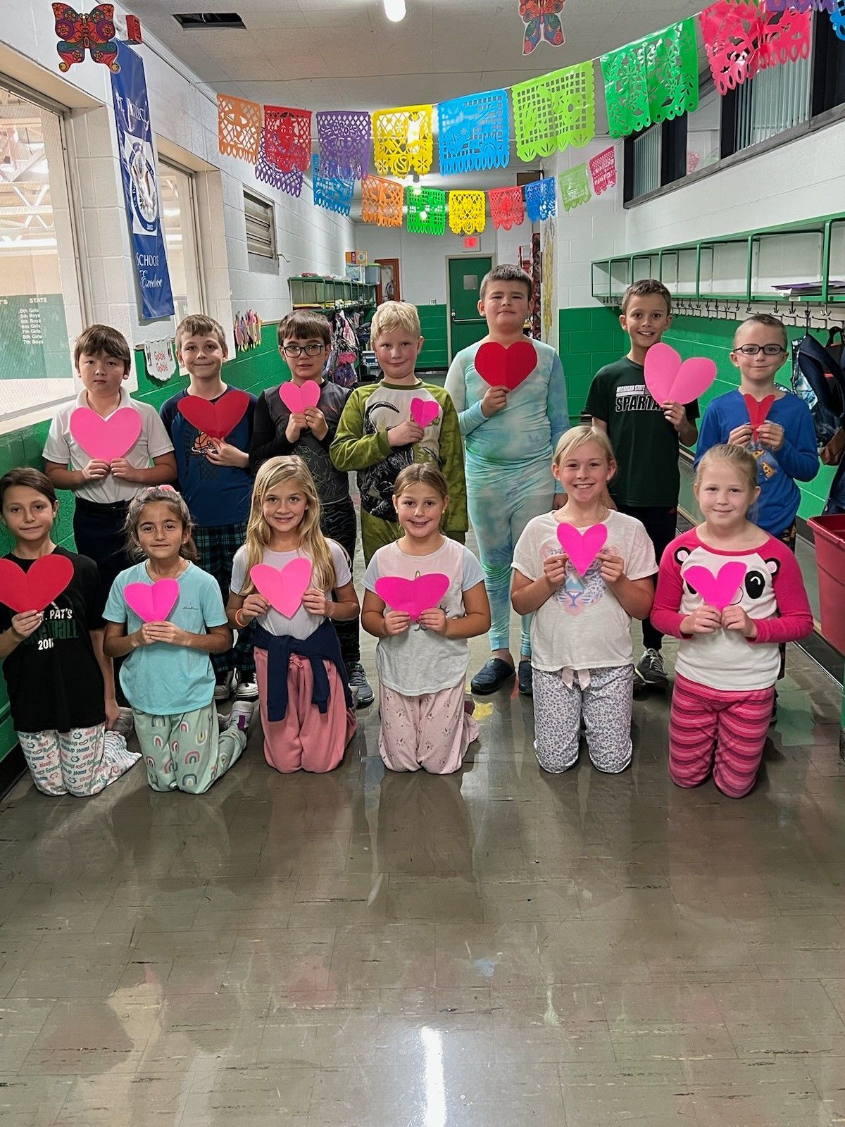 Group of children holding heart-shaped paper, in a school hallway, decorated with colorful flags.