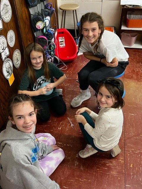 Four young girls seated in a circle, smiling, in a classroom.