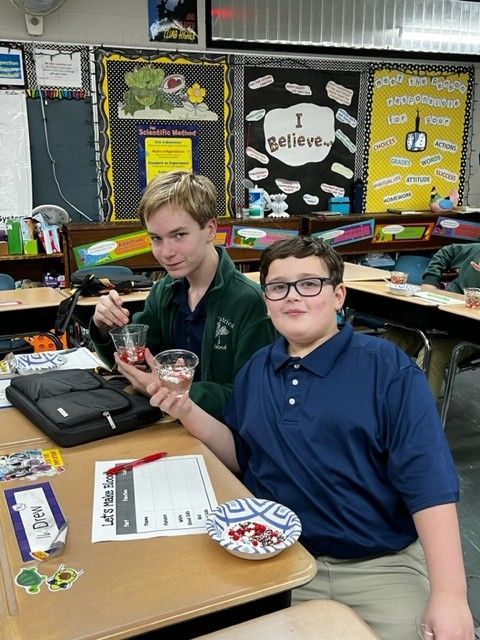 Two students in a classroom with snacks; one smiles, other looks toward camera.