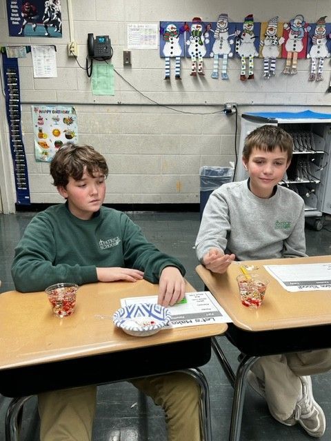 Two boys in a classroom, seated at desks. One holds a treat, the other gestures. Snowman decor on wall.
