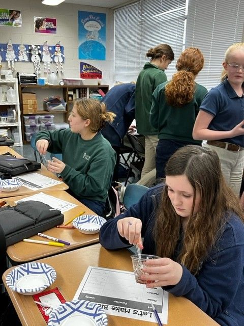 Students in a classroom do a science experiment. They stir liquids in clear beakers, wearing school uniforms.