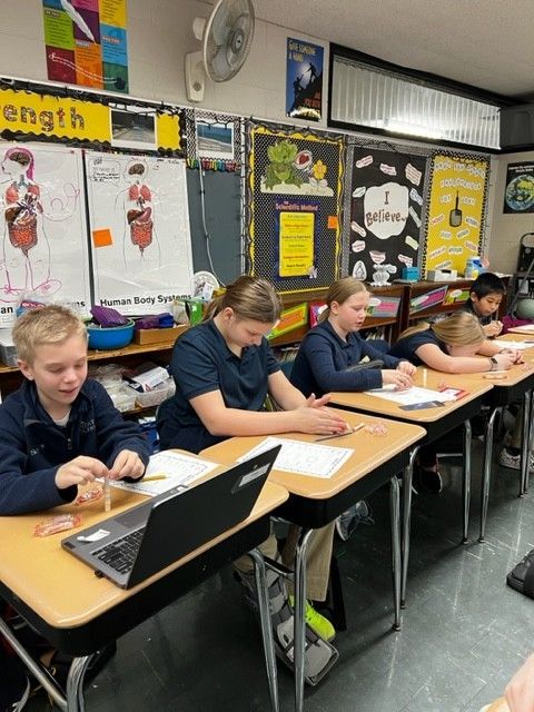 Students in a classroom are seated at desks, working. One has a laptop open.