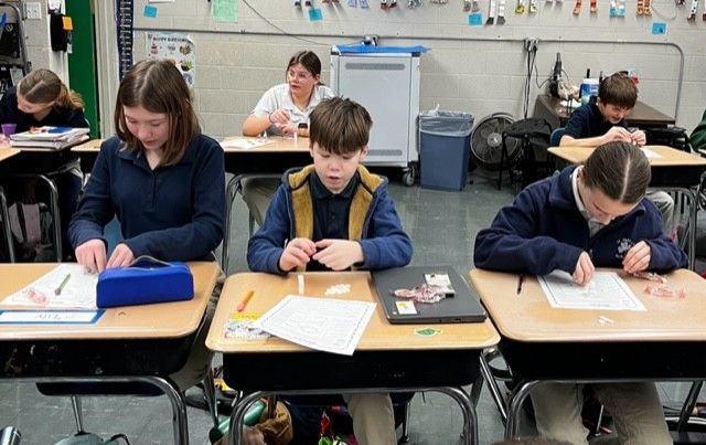 Students in uniforms work at desks in a classroom, focused on tasks, with various school supplies.