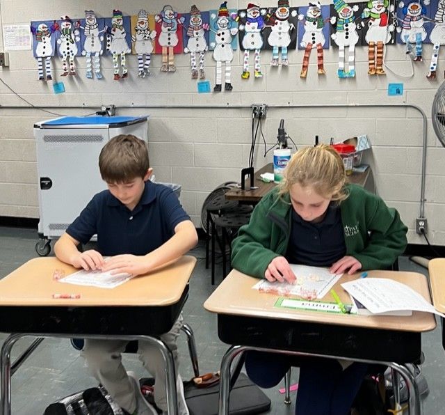 Two students seated at desks, working on paper. Snowman decorations on the wall.