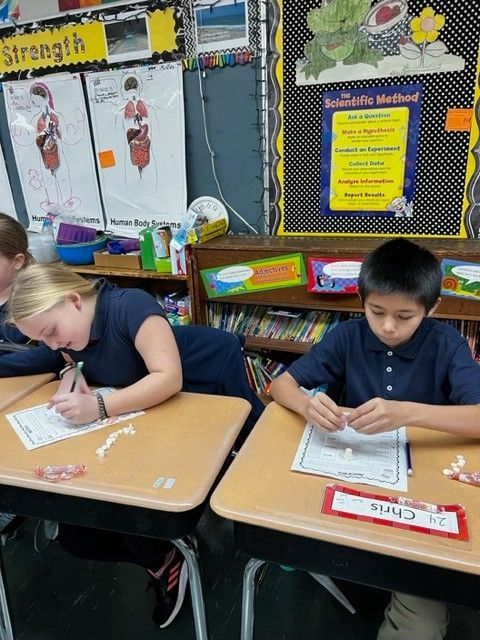 Two students in blue shirts seated at desks, working on assignments. Classroom setting.