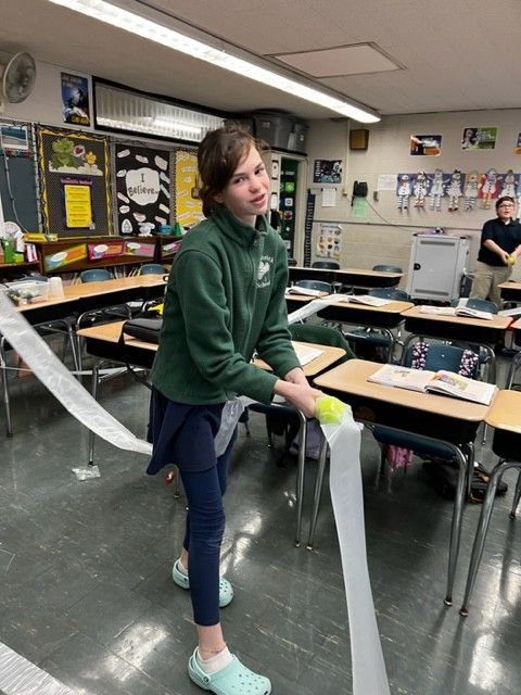 Teenager in green sweatshirt holding a roll of paper in a classroom, smiling at the camera.