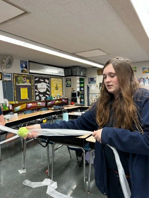 Teenage girl in classroom holds tennis ball and tape, possibly for a physics experiment.