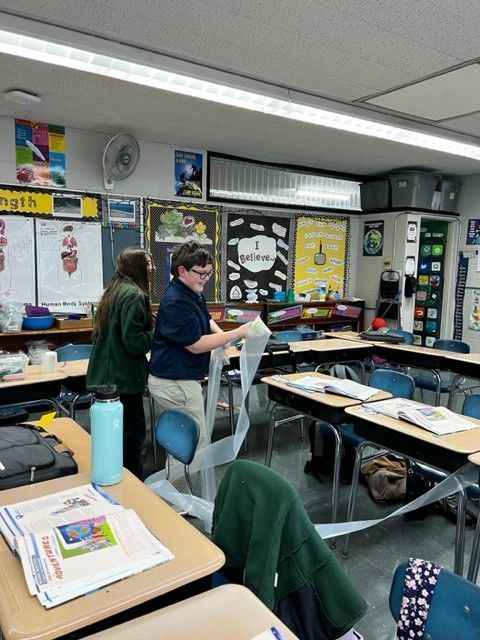 A student smiles, holding paper streamers in a classroom, while another student stands nearby.