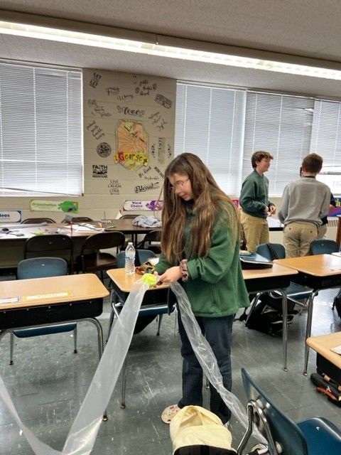 A student in a classroom cuts plastic with scissors, other students in the background.
