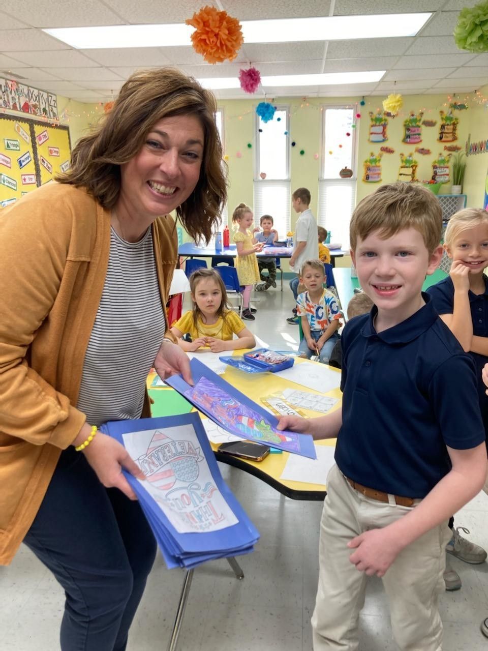 Woman and boy in classroom hold artwork, smiling. Other students and colorful decorations are in the background.