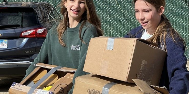 Two young women carrying cardboard boxes outdoors. One smiles, the other looks ahead.