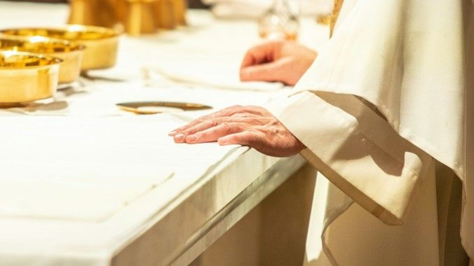 Priest's hands on the altar with chalices in the background during a religious ceremony.