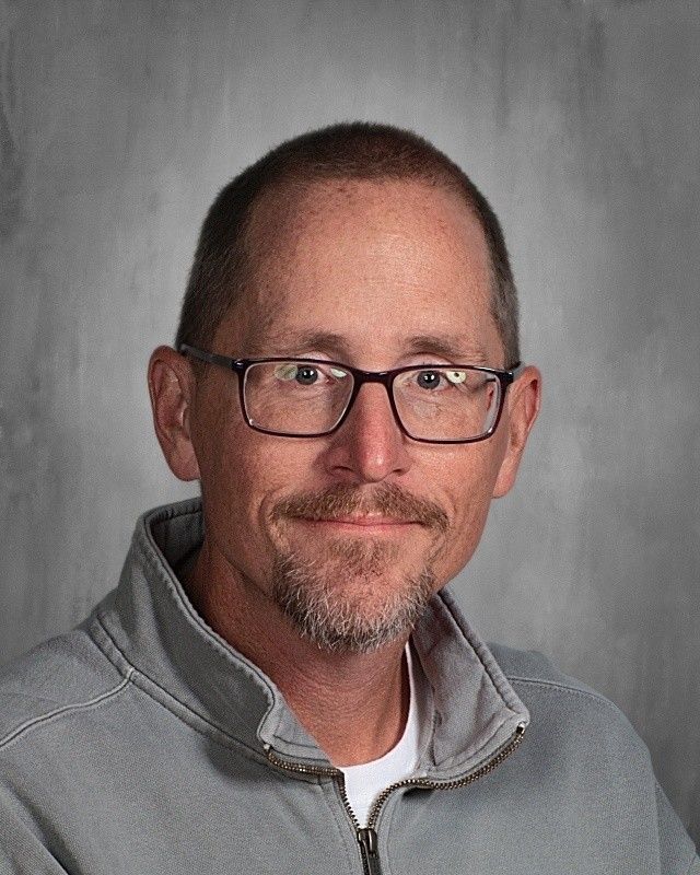 Man with glasses and stubble, wearing a gray zip-up jacket, smiling at the camera.