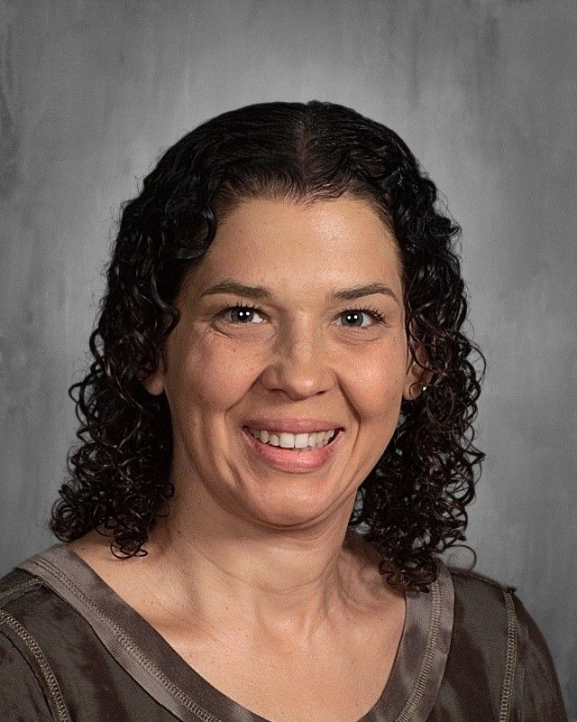 Smiling woman with dark curly hair, wearing a brown top, against a gray backdrop.