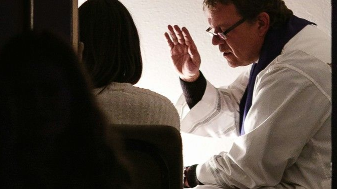 Priest blessing a person sitting in a booth, inside a church.