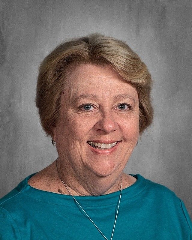 Smiling woman with short blonde hair, wearing a teal top, against a gray backdrop.