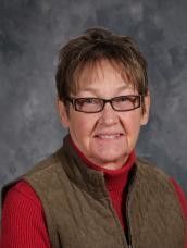 Woman with short brown hair and glasses, wearing a red turtleneck and brown vest.