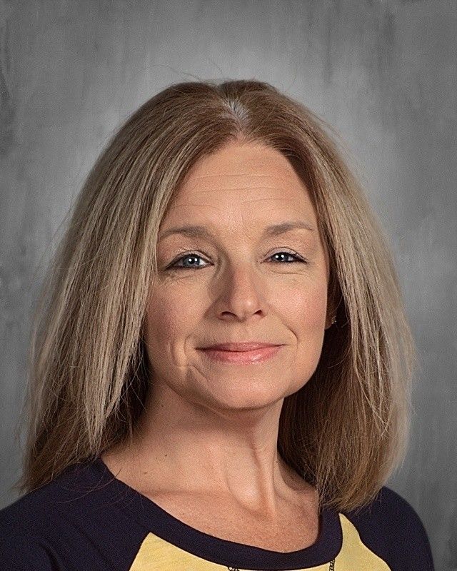 Blonde woman with a neutral expression wearing a navy blue and yellow top. Gray background.