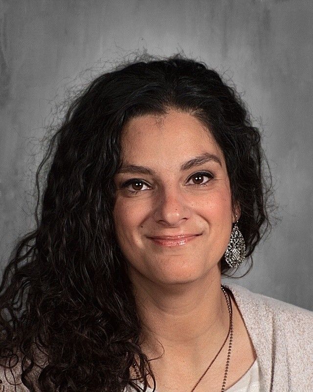 Woman with curly dark hair smiles, wearing earrings and a beige sweater.