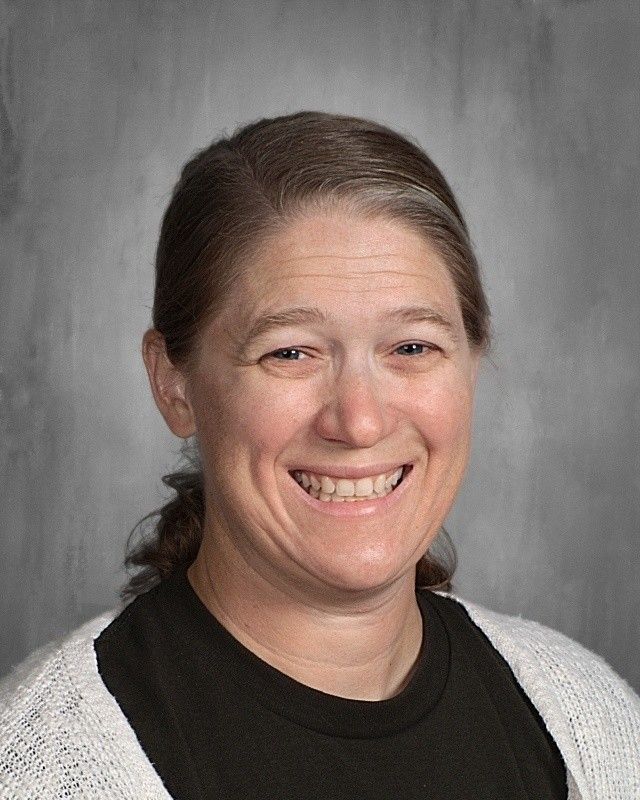 Woman with fair skin, smiling, wearing a black top and white sweater, against a gray backdrop.