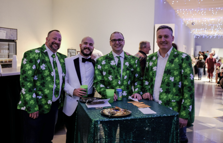 Four men at a St. Patrick's Day event, three in shamrock suits, one in a tuxedo, at a table with green decor.