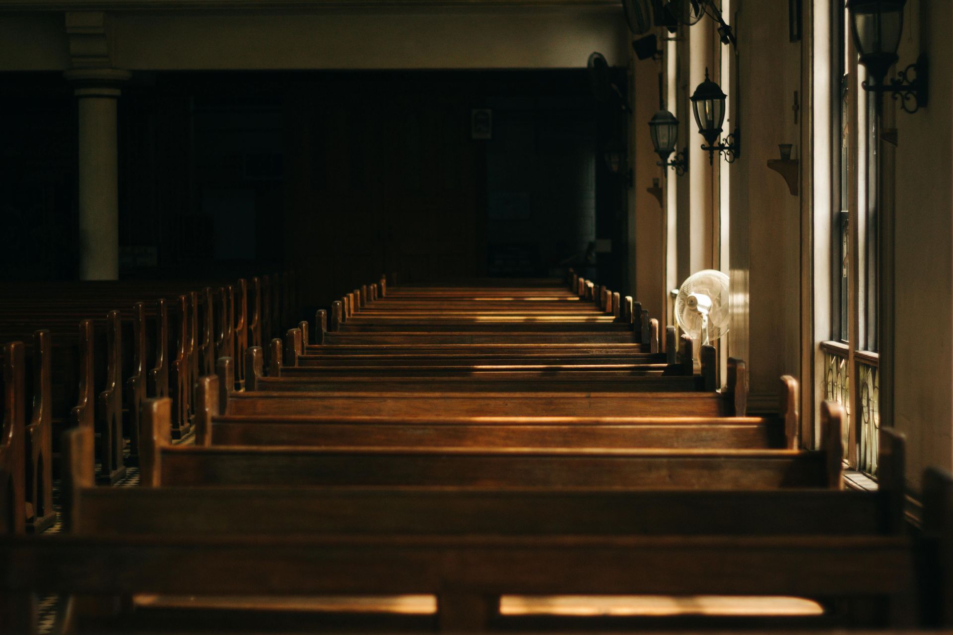 Rows of wooden church pews, bathed in sunlight from a side window, leading towards a dark altar.