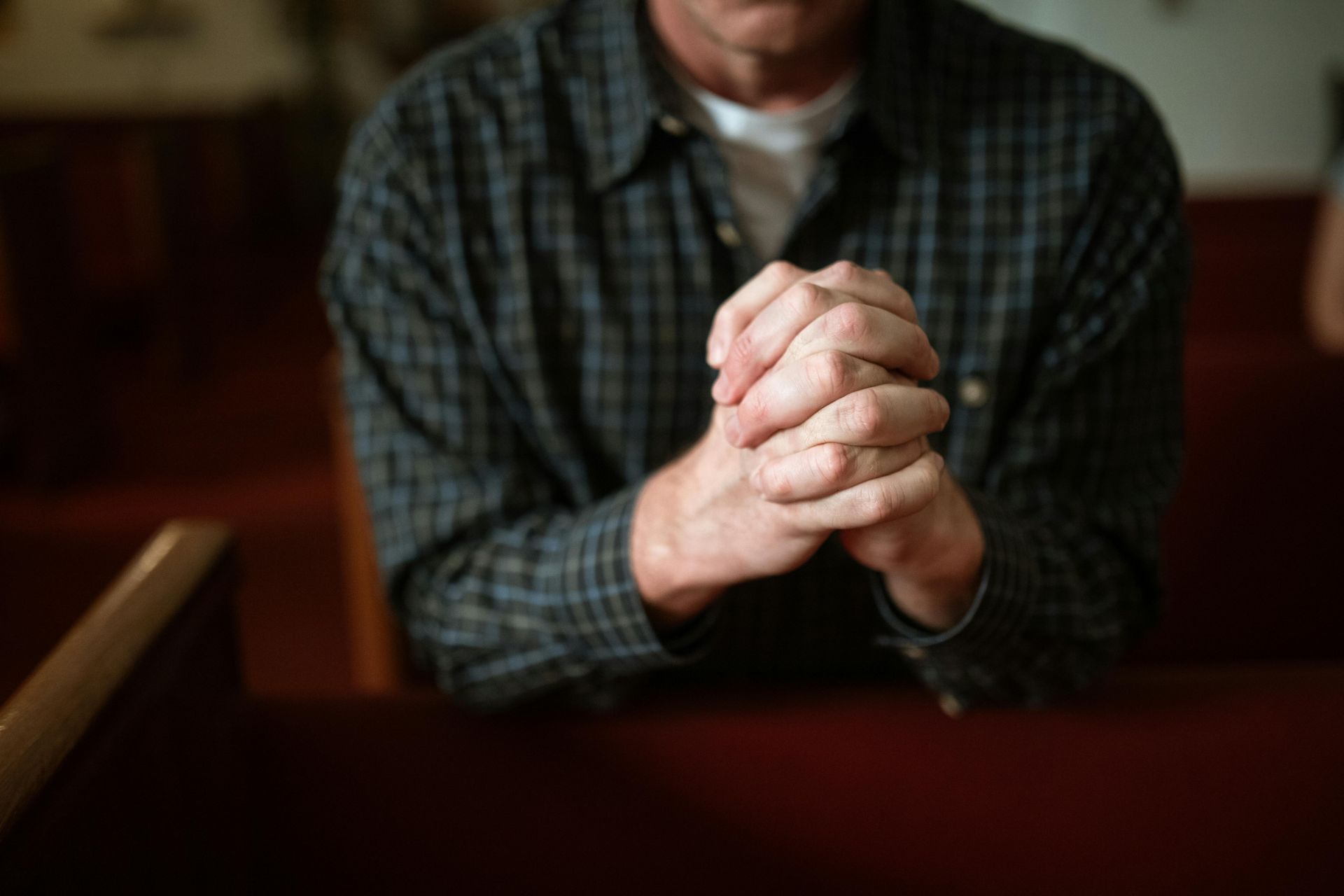 Man in plaid shirt, hands clasped in prayer at a church, resting on pew.