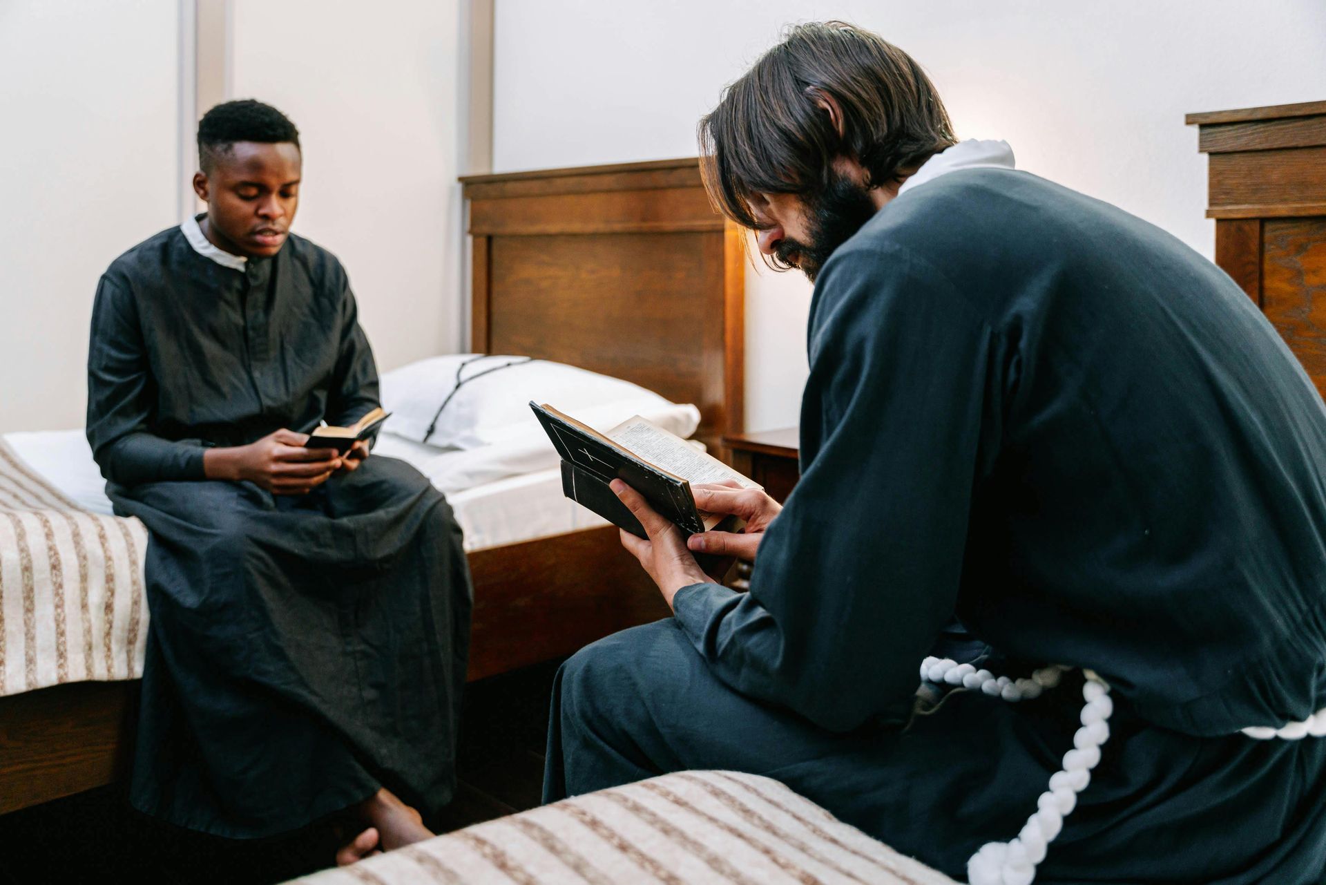 Two people, likely priests, read books in a simple bedroom. One sits on a bed, other kneels.