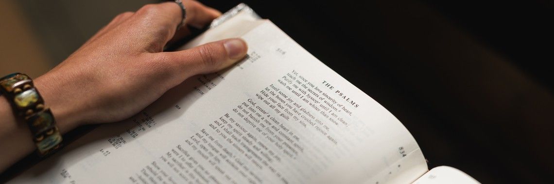 A hand holding open a book with text, wearing a bracelet and a ring, against a dark background.