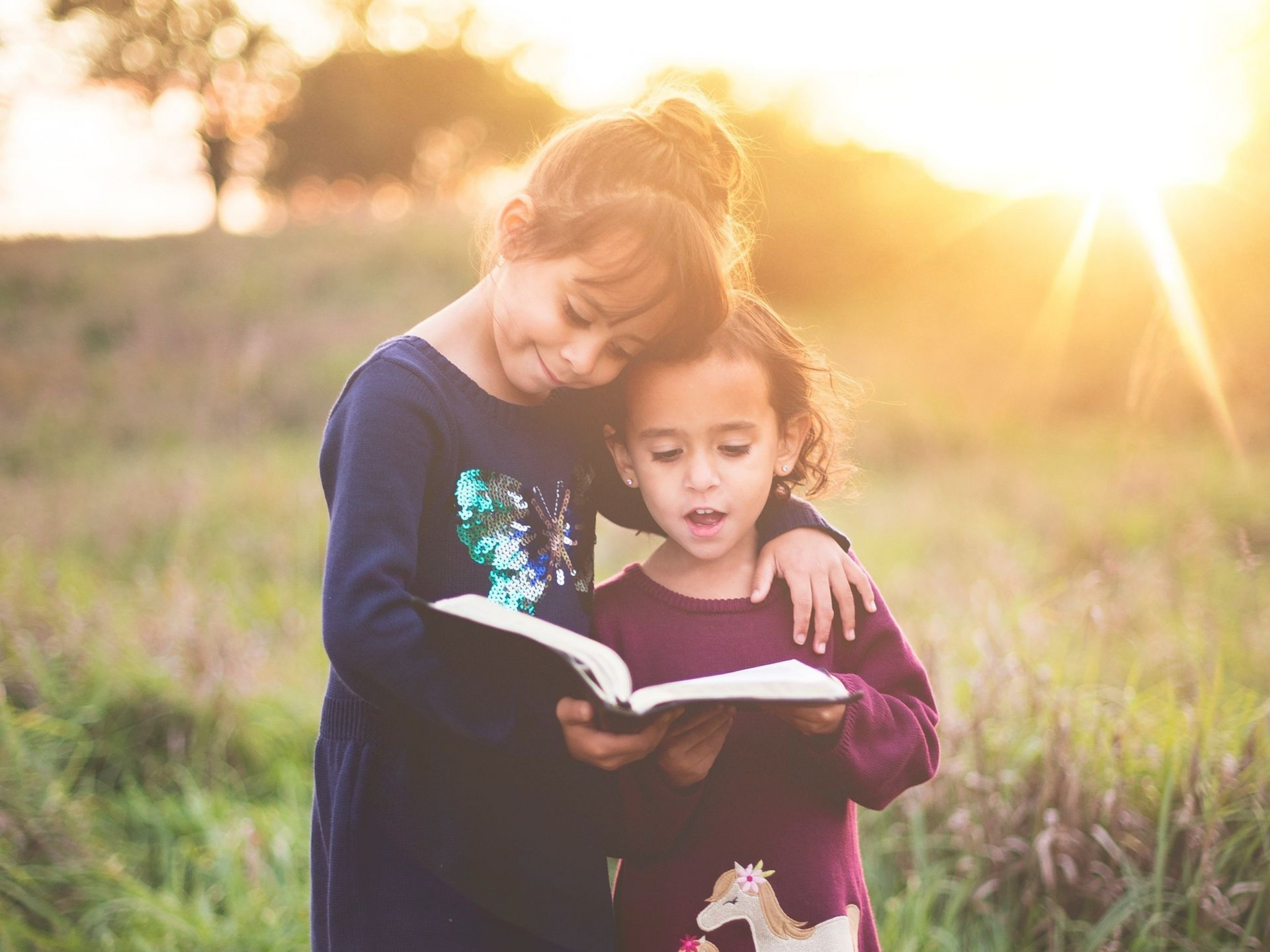 Two young girls reading a book together outdoors in golden sunlight.
