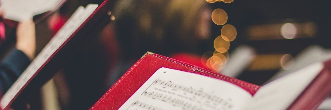 Choir members reading sheet music in a church. Red covers and blurred golden lights in the background.