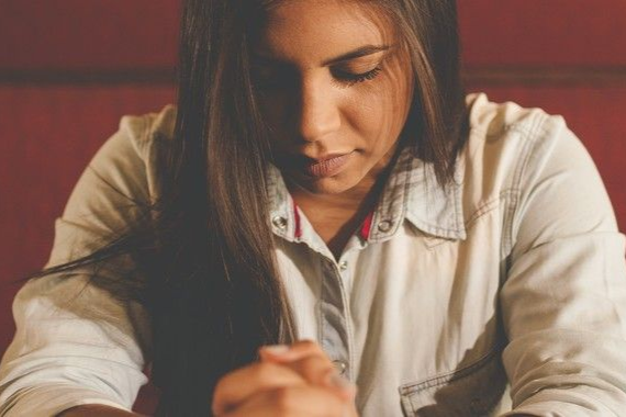 Woman with clasped hands and closed eyes, indoors, possibly praying.