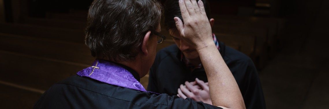 A priest blesses a person in a church. The priest is wearing a purple collar.