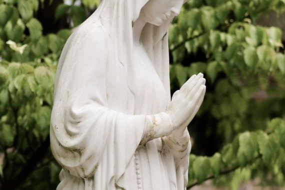 Statue of a praying woman in white robes, hands clasped, head bowed. Green foliage background.