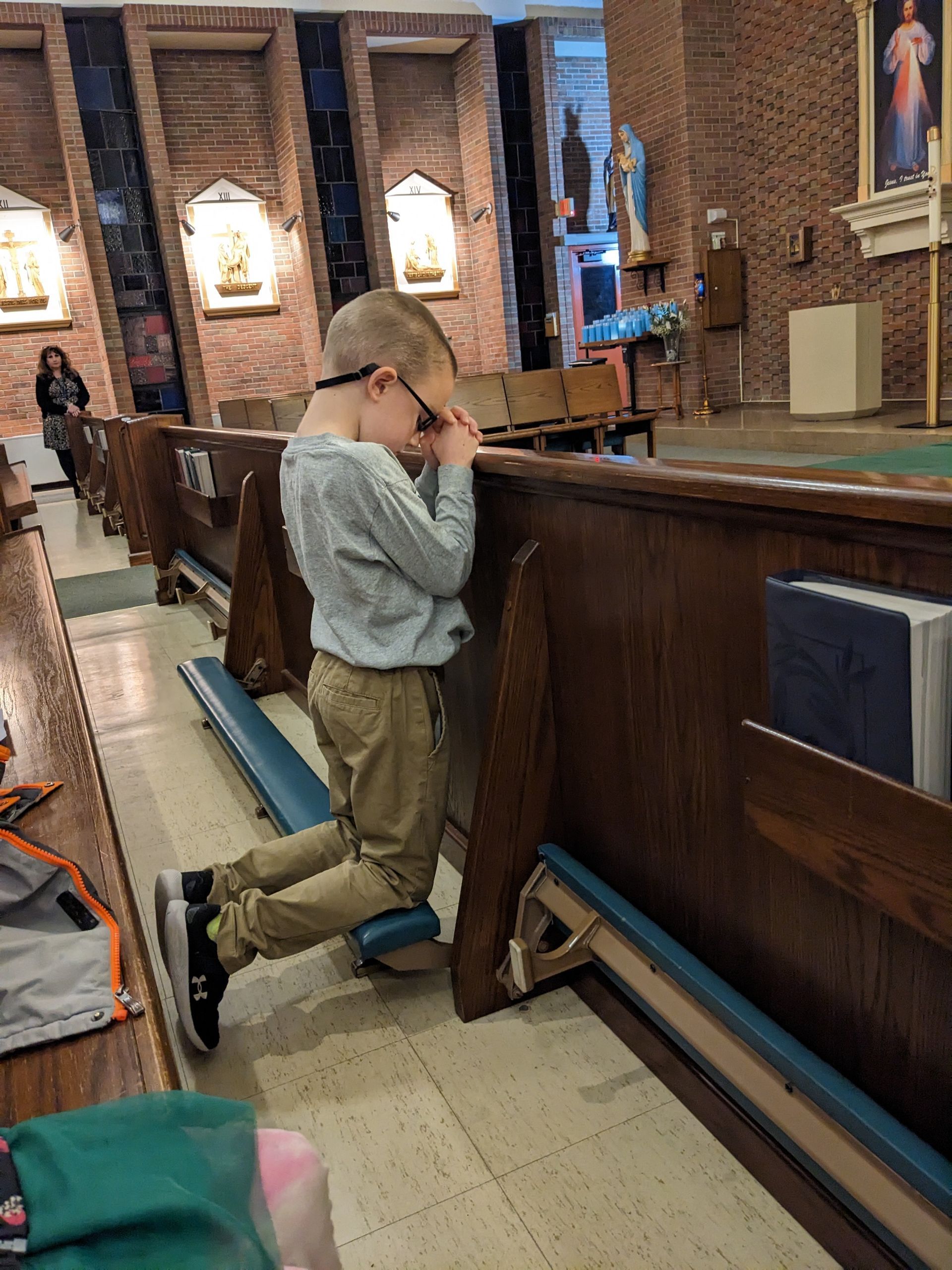 Boy kneeling in church, praying with hands clasped, wearing glasses, khaki pants, and gray shirt.