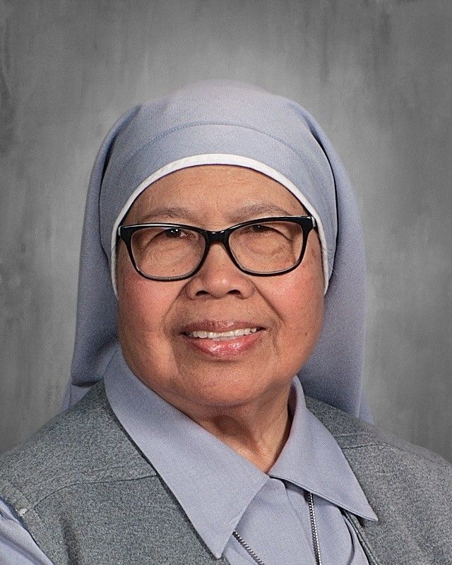 Nun with glasses smiles, wearing a gray habit and collar, against a gray background.