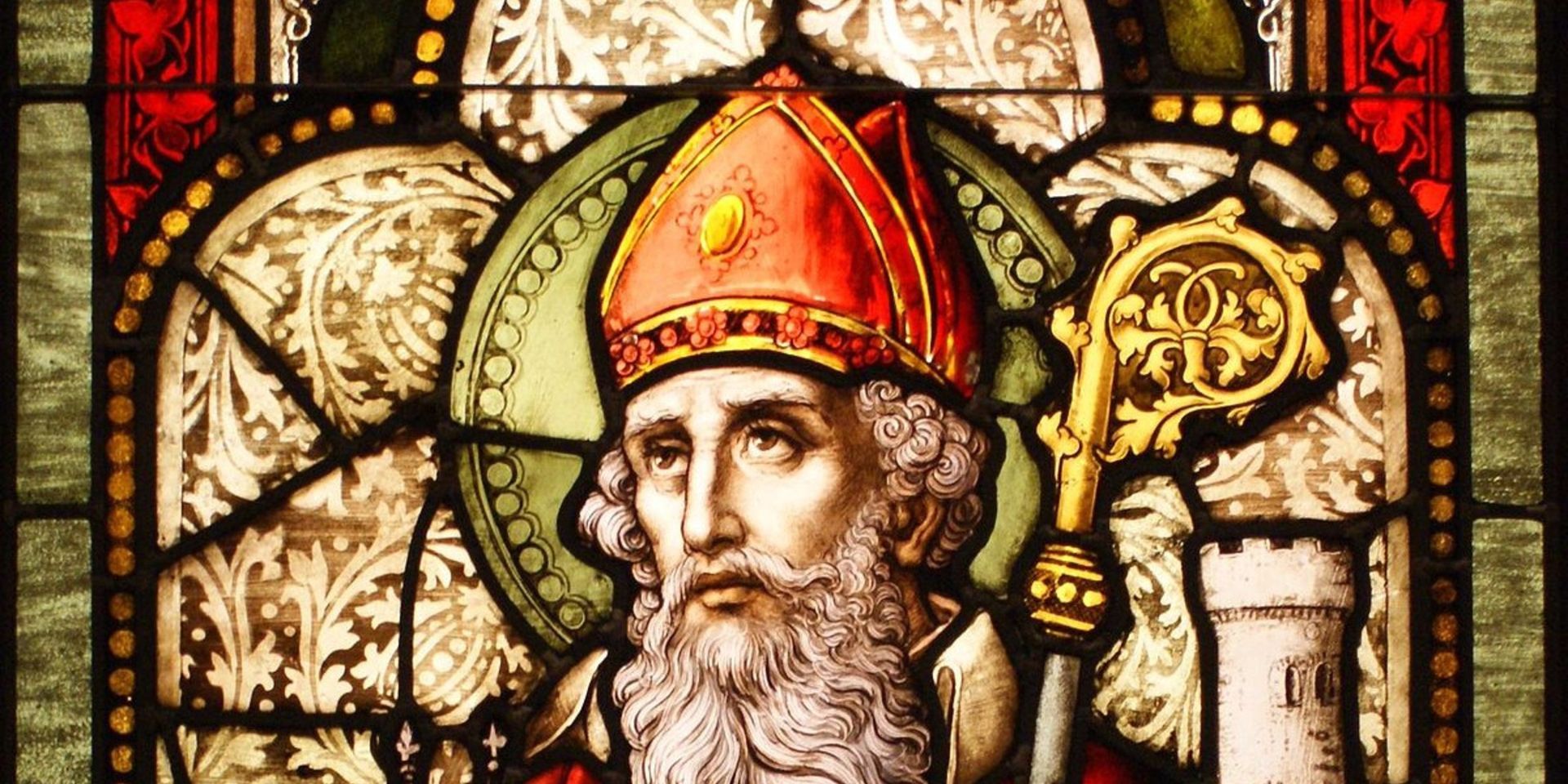 Stained-glass portrait of a bearded man in a red bishop's hat, holding a gold staff, within a church.