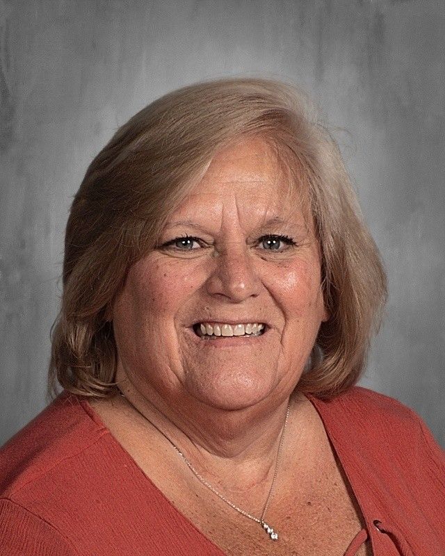 Woman with blonde hair and red shirt smiles, wearing a necklace. Background is grey.