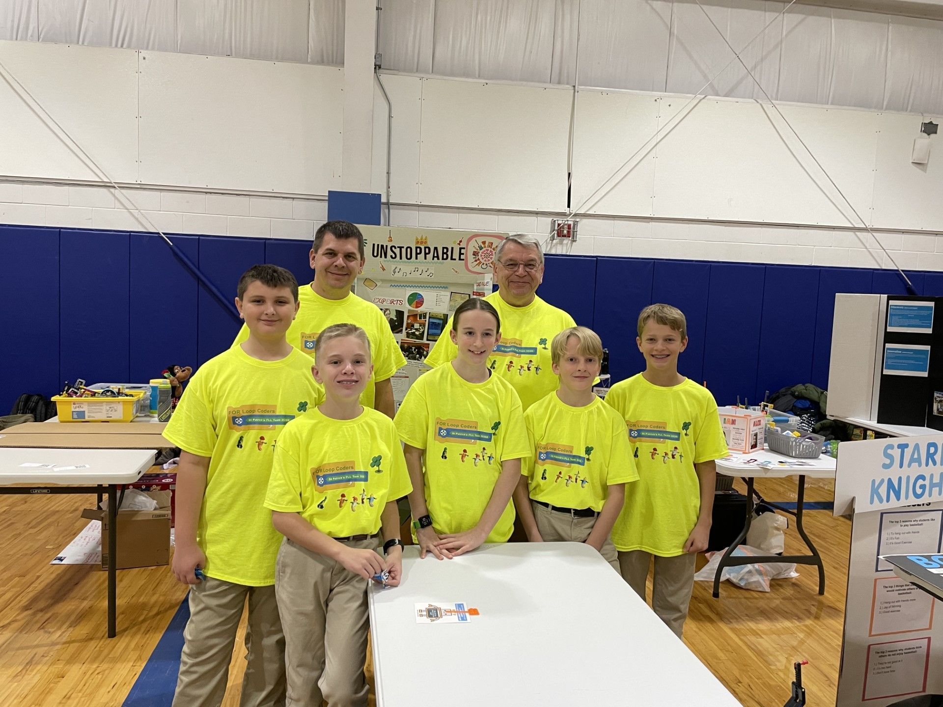 Robotics team with two adults wearing bright yellow shirts, smiling, standing at a table, inside.