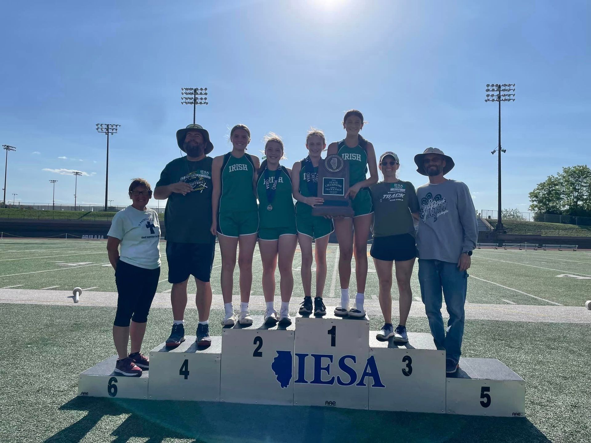 Track team on podium, holding trophy, celebrating victory at a sunny outdoor stadium.