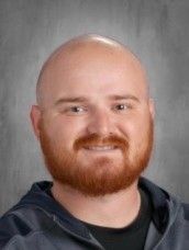 Bald man with a red beard smiling. Wearing a dark shirt, grey zip-up jacket.