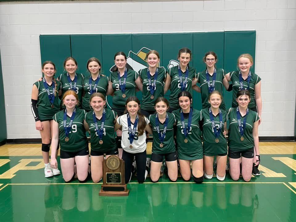 Volleyball team in green jerseys kneels with trophy; green and white wall.