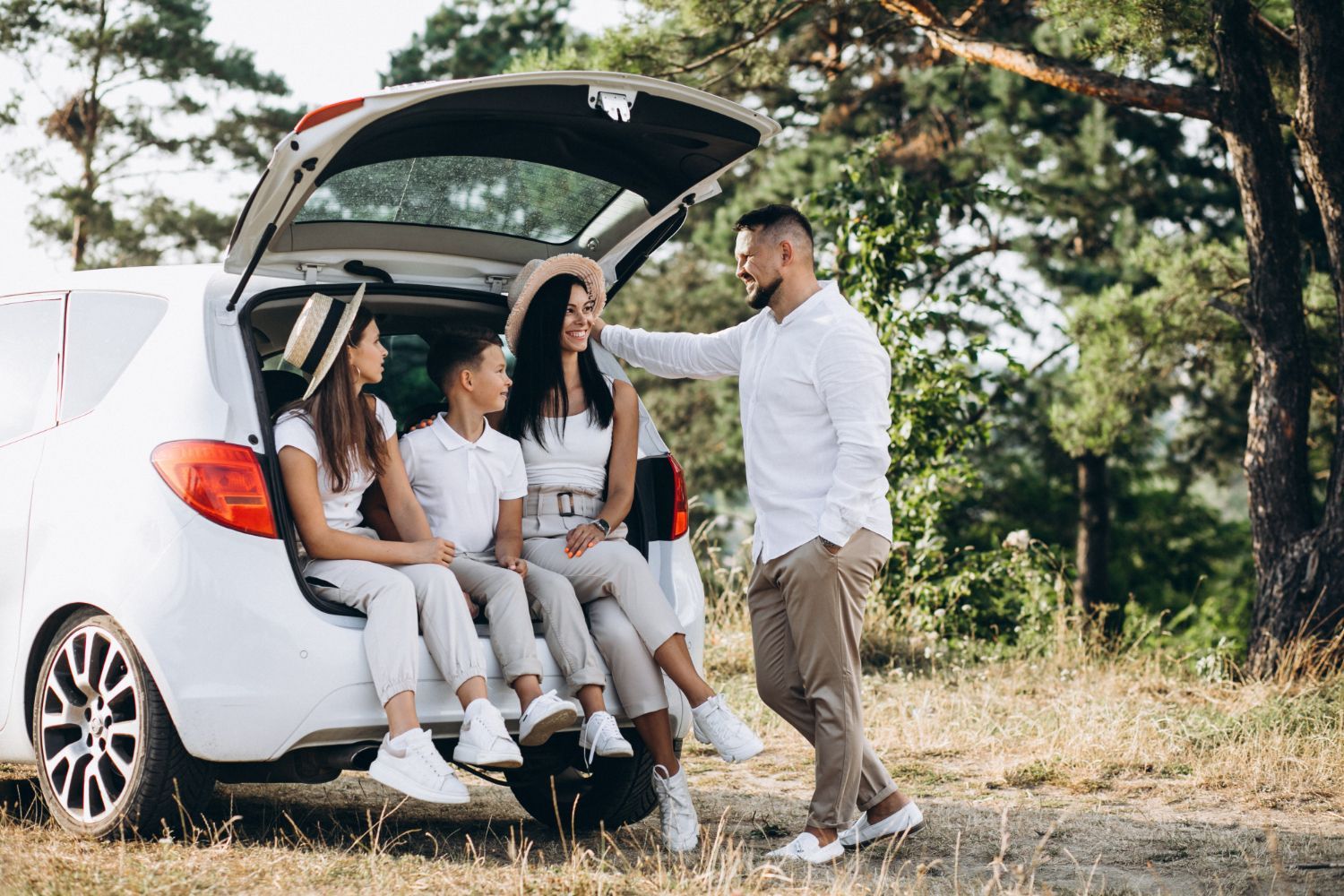 Family of four sitting in an open car trunk, man standing beside them outdoors.